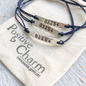 Pode incluir: Tr&ecirc;s pulseiras de cord&atilde;o azul com pingentes de prata que dizem "03254". As pulseiras est&atilde;o em uma bolsa de tecido branco com as palavras "Positive Charm Handmade Jewelry" impressas nela.