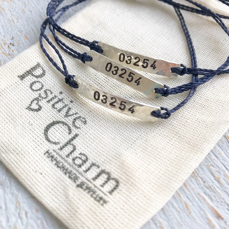 Pode incluir: Tr&ecirc;s pulseiras de cord&atilde;o azul com pingentes de prata que dizem "03254". As pulseiras est&atilde;o em uma bolsa de tecido branco com as palavras "Positive Charm Handmade Jewelry" impressas nela.