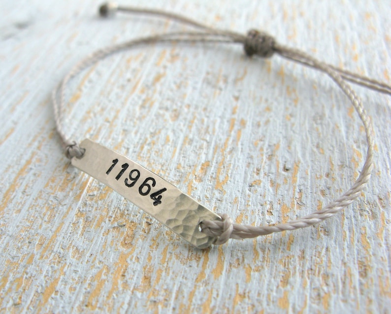 Pode incluir: Um bracelete de corda cinzenta com uma etiqueta prateada que diz "11964".