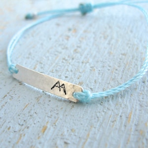 May include: A light blue braided cord bracelet with a silver charm featuring a mountain silhouette.