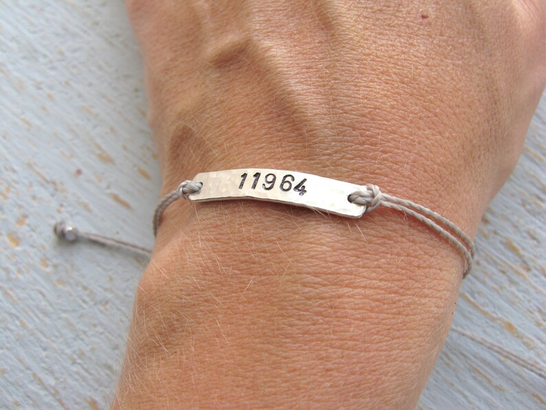 Pode incluir: Uma pulseira de prata com um cord&atilde;o cinza e uma etiqueta de prata que diz "11964".