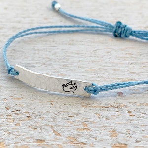 May include: A light blue adjustable bracelet with a hammered silver bar. The bar features a black outline of a dove. The bracelet is made of blue cord and has a sliding knot closure.