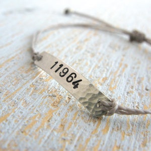 Pode incluir: Um bracelete de corda cinzenta com uma etiqueta prateada que diz "11964".