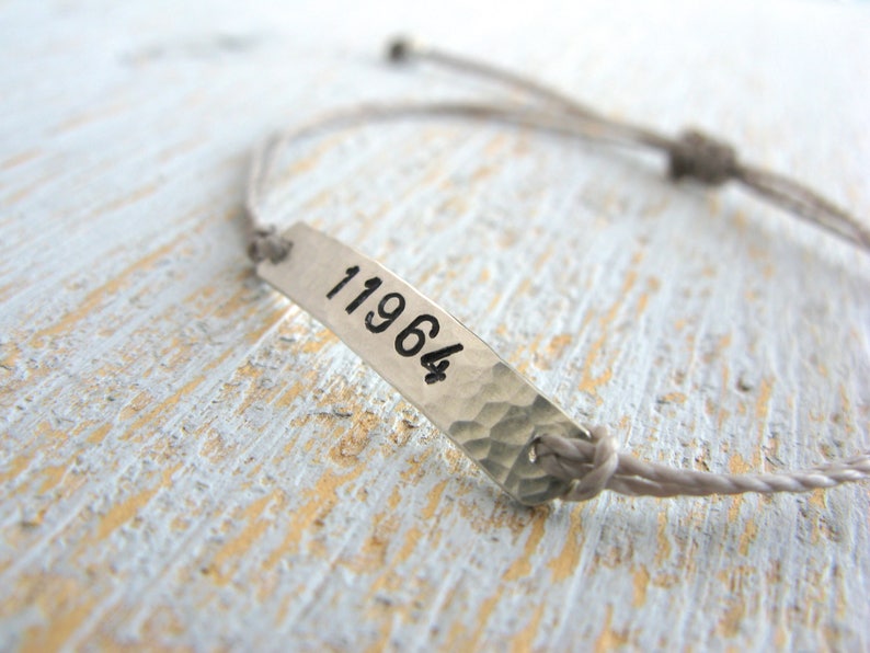 Pode incluir: Um bracelete de corda cinzenta com uma etiqueta prateada que diz "11964".
