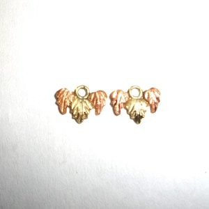 May include: Two small, gold-toned leaf charms with a three-leaf design. The leaves have a mix of gold and copper tones, with a small loop at the top for attaching to jewelry. The charms are set against a white background.