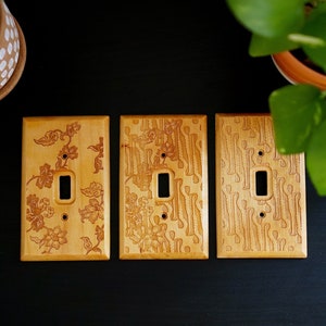 May include: Three wooden light switch covers with different engraved patterns. The covers are light brown and have a natural wood finish.
