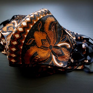 May include: A stack of black and orange fabric face masks with a floral pattern. The masks have black elastic ear loops.