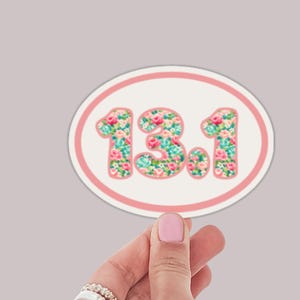 May include: Oval sticker with a pink border and the text "13.1" in a floral pattern of pink, blue, and green. The sticker is held up by a hand with a pink painted nail.