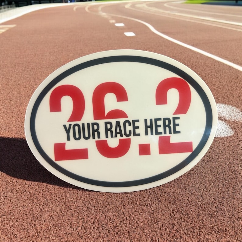 Puede incluir: Letrero ovalado con el texto "26.2 YOUR RACE HERE" en rojo y negro. El letrero tiene un fondo color crema con un borde negro y blanco. El letrero est&aacute; sobre una pista de atletismo roja.
