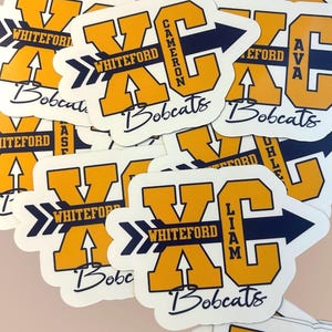 May include: A collection of die-cut stickers featuring a large yellow letter X and C with a dark blue arrow pointing right. The text Whiteford is printed on the arrow, and the word Bobcats is written in script below. Individual names like Cameron, Ava, and Liam are printed vertically on the letter C.