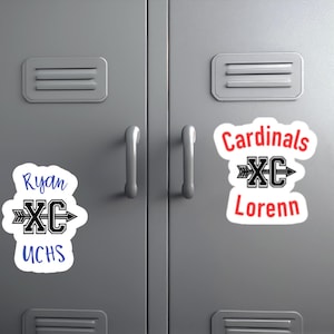 May include: Two die-cut vinyl stickers applied to a gray metal locker. The left sticker features the blue text Ryan and UCHS with a black arrow and XC logo. The right sticker features the red text Cardinals and Lorenn with a black arrow and XC logo.