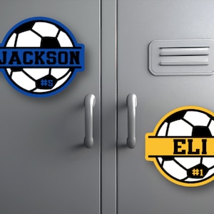 Puede incluir: Dos calcomanías con temática de balón de fútbol con texto personalizado. Una es azul con "JACKSON" y "#5", la otra es amarilla con "ELI" y "#1". Ambas están sobre una superficie metálica gris.