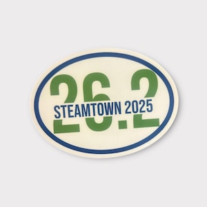 Puede incluir: Adhesivo ovalado con el texto "26.2 STEAMTOWN 2025" en verde y azul. El adhesivo tiene un fondo crema con un borde azul y verde. El dise&ntilde;o es simple y audaz, para corredores de marat&oacute;n.