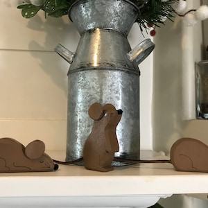 May include: Three wooden mouse figurines on a white shelf. The mice are brown and have black eyes and noses. One mouse is standing upright, while the other two are lying down. The mice are in front of a galvanized metal pitcher with a floral arrangement.