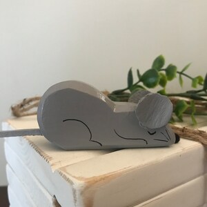 May include: A gray wooden mouse figurine with a painted face and closed eyes. The mouse is lying on a stack of white wooden boxes.