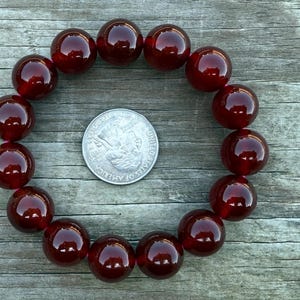 May include: A dark red beaded bracelet with large, round beads. The bracelet is arranged in a circle, with a quarter coin in the center for size comparison. The beads have a glossy finish and reflect light.