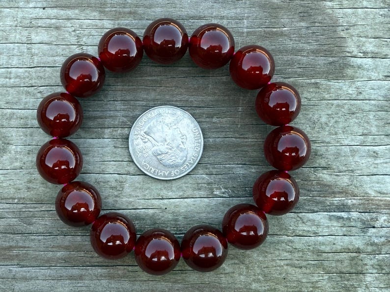 May include: A dark red beaded bracelet with large, round beads. The bracelet is arranged in a circle, with a quarter coin in the center for size comparison. The beads have a glossy finish and reflect light.