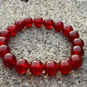 May include: A bracelet composed of large, round, translucent red beads. The beads are strung together to form a circular shape, resting on a textured, gray surface. The beads have a glossy finish, reflecting light.