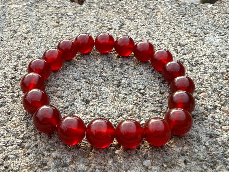 May include: A bracelet composed of large, round, translucent red beads. The beads are strung together to form a circular shape, resting on a textured, gray surface. The beads have a glossy finish, reflecting light.