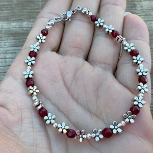 May include: A delicate bracelet featuring alternating dark red beads and silver-colored flower-shaped charms. The bracelet is secured with a silver clasp. This jewelry piece is suitable for everyday wear or special occasions.