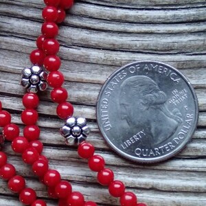 Puede incluir: Primer plano de un collar de cuentas de coral rojo con detalles de flores plateadas. El collar est&aacute; colocado sobre una superficie de madera junto a una moneda de 25 centavos de d&oacute;lar estadounidense.