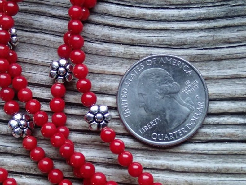 Puede incluir: Primer plano de un collar de cuentas de coral rojo con detalles de flores plateadas. El collar est&aacute; colocado sobre una superficie de madera junto a una moneda de 25 centavos de d&oacute;lar estadounidense.