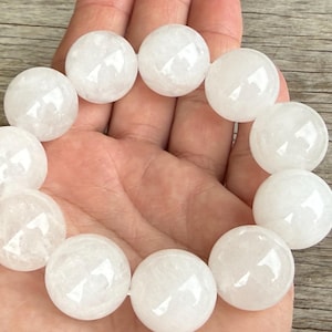 May include: A white quartz crystal bead bracelet. The bracelet features large, round, polished beads. The beads are a translucent white color. The bracelet is held in a hand.