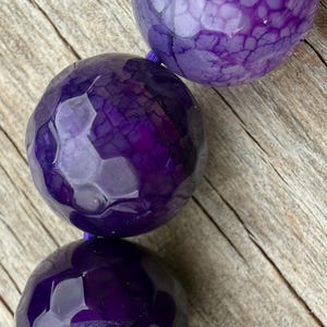 May include: Close-up of a strand of large, faceted, deep purple beads. The beads have a textured surface and are strung on a thin, purple cord. The background is a light brown, wooden surface.
