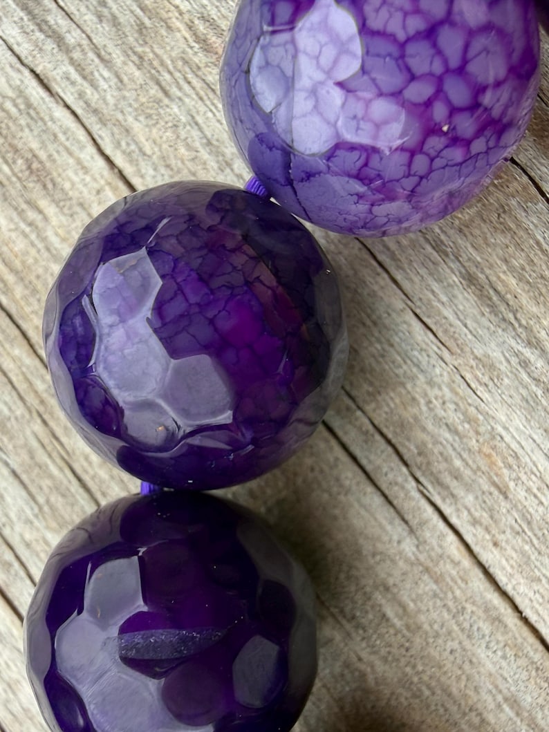 May include: Close-up of a strand of large, faceted, deep purple beads. The beads have a textured surface and are strung on a thin, purple cord. The background is a light brown, wooden surface.