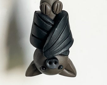 May include: A handcrafted bat-shaped charm with a dark brown body and black wings, hanging upside down. The bat has small, round eyes and a detailed face. A light green bead is at the top, connected by a silver-colored metal loop.