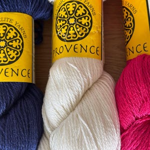 May include: Close-up of three skeins of Classic Elite Yarns in navy blue, white, and hot pink. Each skein has a yellow label with the word "Provence" and a decorative emblem. The yarn is tightly wound and appears soft.
