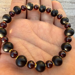 May include: A beaded bracelet featuring alternating matte black and deep red beads. The bracelet's design includes larger, rounded black beads interspersed with smaller, round red beads, all connected by gold-colored accents. This piece is a stylish accessory.
