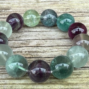 May include: A bracelet featuring large, round beads in shades of green, purple, and clear. The beads have a polished, reflective surface and are strung together. The bracelet is displayed on a wooden surface.