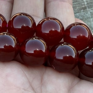 May include: A strand of large, polished, round beads in a deep burgundy color. The beads have a glossy, reflective surface, and are held in a person's hand. The beads are evenly sized and appear to be strung together.