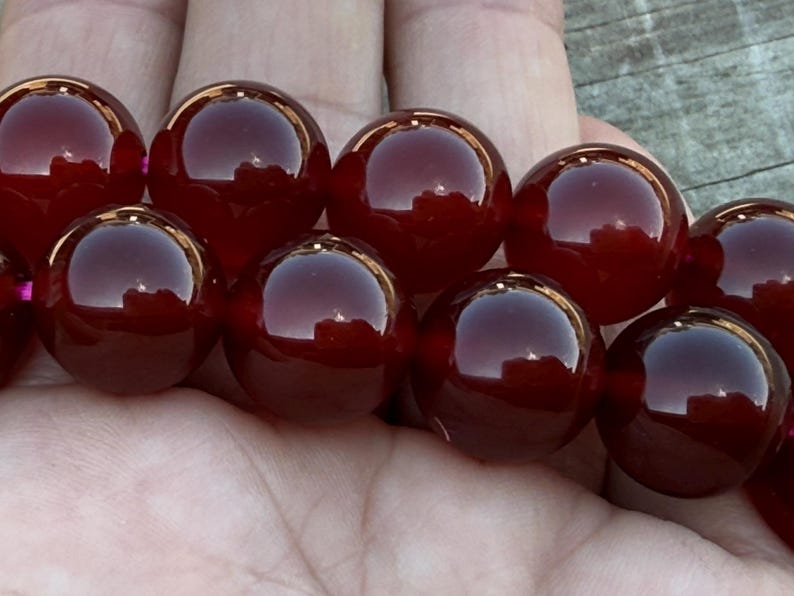 May include: A strand of large, polished, round beads in a deep burgundy color. The beads have a glossy, reflective surface, and are held in a person's hand. The beads are evenly sized and appear to be strung together.