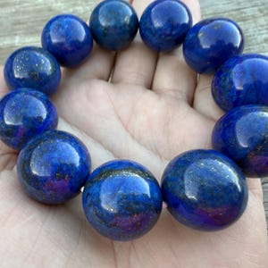 May include: A stretch bracelet composed of large, round, polished lapis lazuli beads. The beads feature a deep royal blue color with visible natural inclusions of gold-colored pyrite and dark grey mineral veins throughout the smooth, spherical surfaces.