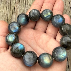 May include: A bracelet of large, round, polished labradorite beads. The beads are primarily gray with iridescent flashes of blue, green, and gold. The bracelet is held in a hand, showcasing the natural variations and colors of the gemstones.