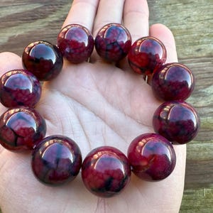 May include: A beaded bracelet featuring large, round beads in shades of deep red and burgundy. The beads have a marbled appearance with dark veins, creating a unique pattern. The bracelet is displayed on a hand.