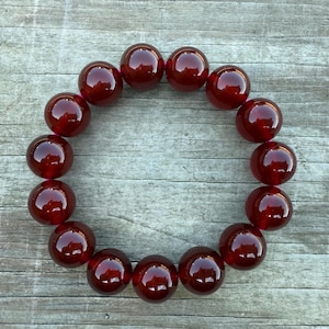 May include: A circular arrangement of large, polished, dark red beads. The beads are evenly spaced, creating a ring shape. The beads have a glossy finish, reflecting light. The background is a weathered, gray wooden surface.