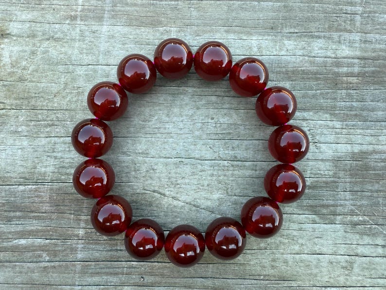 May include: A circular arrangement of large, polished, dark red beads. The beads are evenly spaced, creating a ring shape. The beads have a glossy finish, reflecting light. The background is a weathered, gray wooden surface.