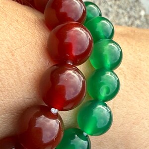 May include: A close-up of two beaded bracelets. One bracelet features large, round, polished beads in a deep red color. The other bracelet has similar beads in a vibrant green. The beads are strung together on a white string.