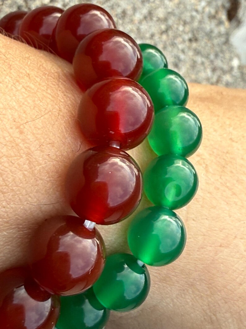 May include: A close-up of two beaded bracelets. One bracelet features large, round, polished beads in a deep red color. The other bracelet has similar beads in a vibrant green. The beads are strung together on a white string.
