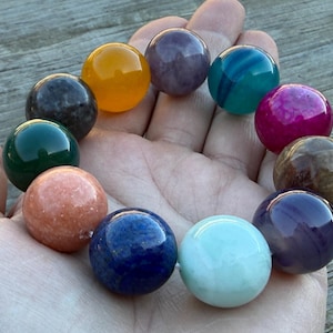 May include: A collection of polished gemstone spheres in various colors, including green, orange, purple, blue, and pink. The spheres are held in a hand, showcasing their smooth, reflective surfaces. The gemstones are likely used for decorative or spiritual purposes.