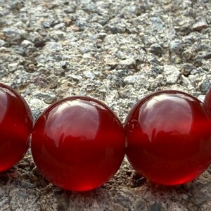 May include: Close-up of a row of large, glossy, red glass beads. The beads are spherical and reflect light, set against a textured, gray stone surface. The beads have a deep, rich color.