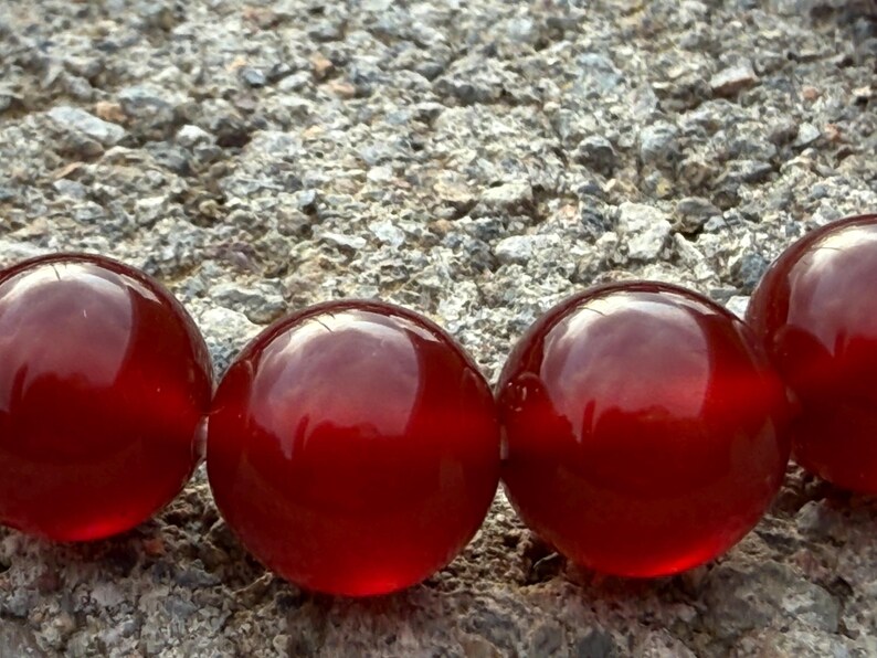 May include: Close-up of a row of large, glossy, red glass beads. The beads are spherical and reflect light, set against a textured, gray stone surface. The beads have a deep, rich color.