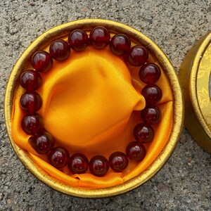 May include: A beaded bracelet with dark red, round beads rests on a gold-colored satin cloth inside a round, gold-colored box. The box is open, revealing the bracelet and cloth.