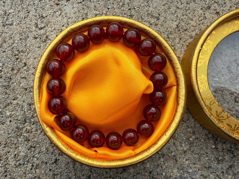 May include: A beaded bracelet with dark red, round beads rests on a gold-colored satin cloth inside a round, gold-colored box. The box is open, revealing the bracelet and cloth.
