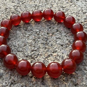 May include: A red beaded bracelet composed of large, round, polished beads. The beads are a deep, translucent red color, strung together to form a circular shape. The bracelet is resting on a textured, gray surface.