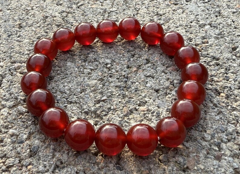 May include: A red beaded bracelet composed of large, round, polished beads. The beads are a deep, translucent red color, strung together to form a circular shape. The bracelet is resting on a textured, gray surface.