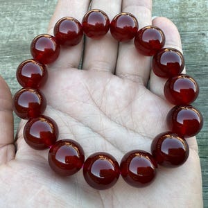 May include: A bracelet featuring large, polished, round beads in a deep, rich red color. The beads are strung together, forming a circular shape. The bracelet is displayed on a hand, showcasing the jewelry's size and color.